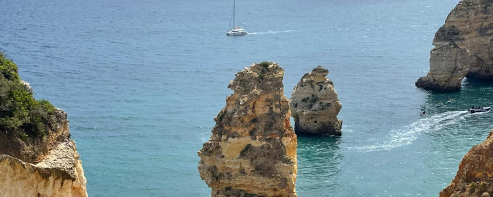 Blick auf das Meer. Man sieht blauen Himmel, Felsen und ein Segelboot.
