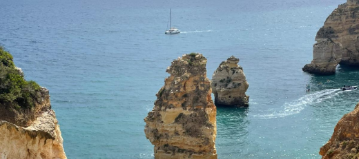 Blick auf das Meer. Man sieht blauen Himmel, Felsen und ein Segelboot.