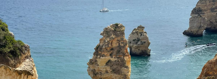 Blick auf das Meer. Man sieht blauen Himmel, Felsen und ein Segelboot.