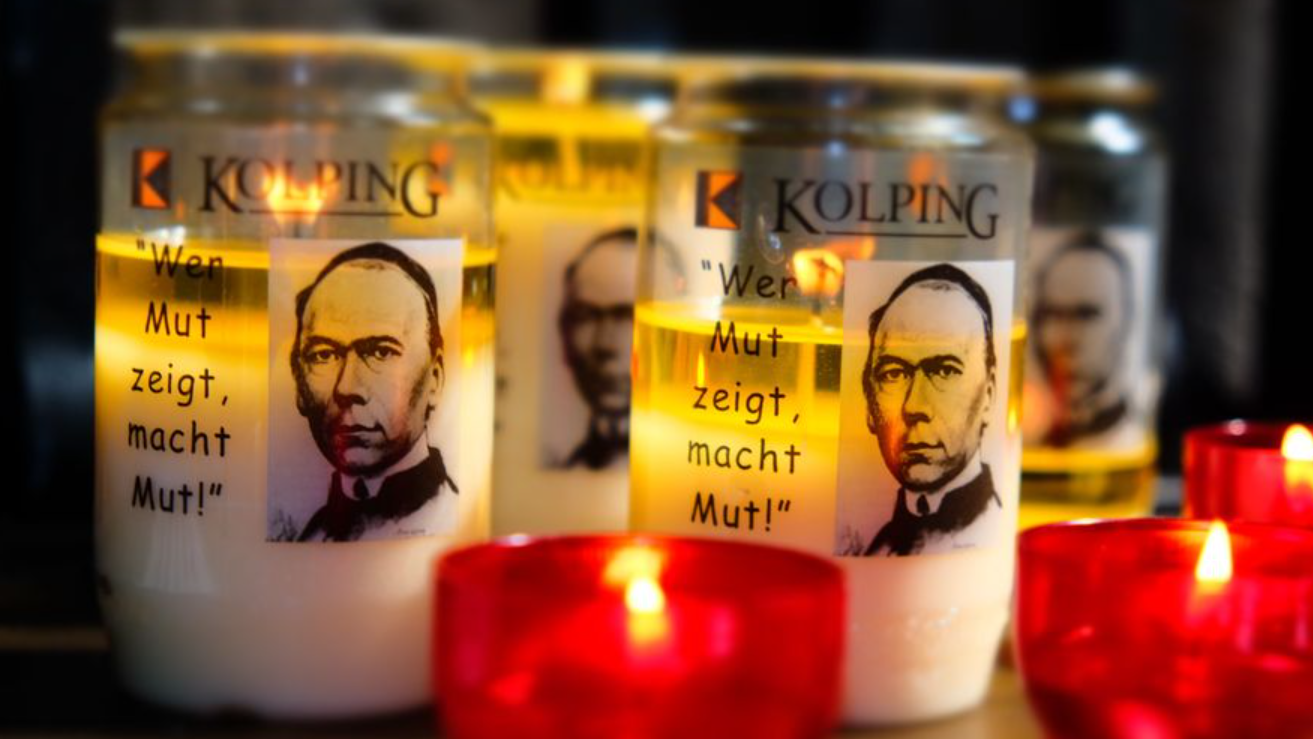 Das Bild zeigt entzündete Kerzen in roten Haltern im Vordergrund, dahinter Öllichter mit dem Portrait Adolph Kolpings.
