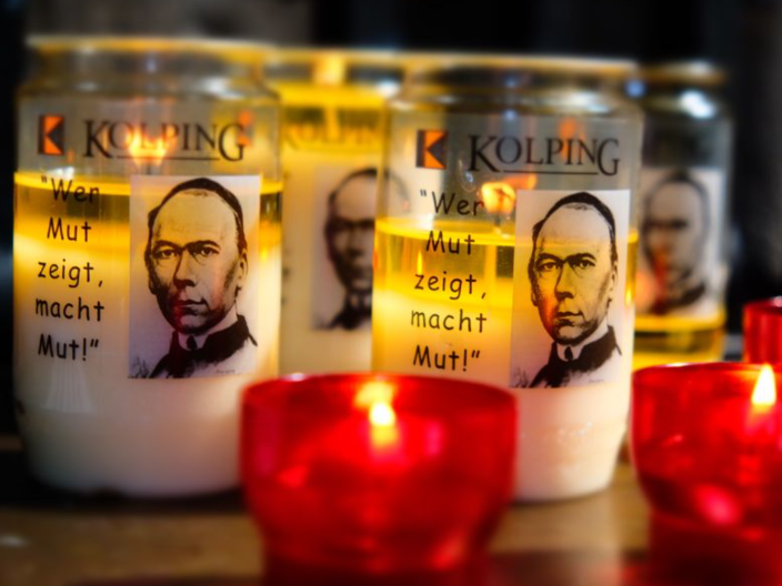 Das Bild zeigt entzündete Kerzen in roten Haltern im Vordergrund, dahinter Öllichter mit dem Portrait Adolph Kolpings.