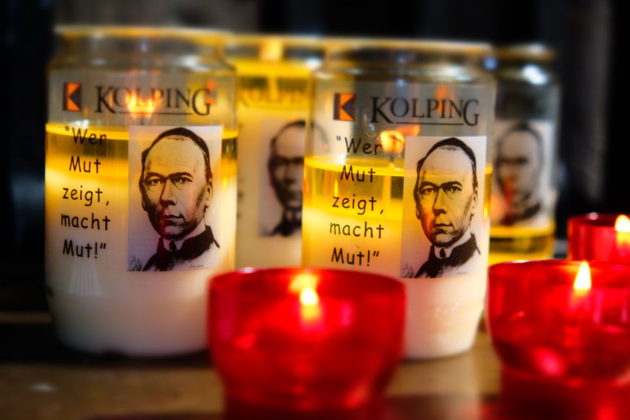 Das Bild zeigt entzündete Kerzen in roten Haltern im Vordergrund, dahinter Öllichter mit dem Portrait Adolph Kolpings.
