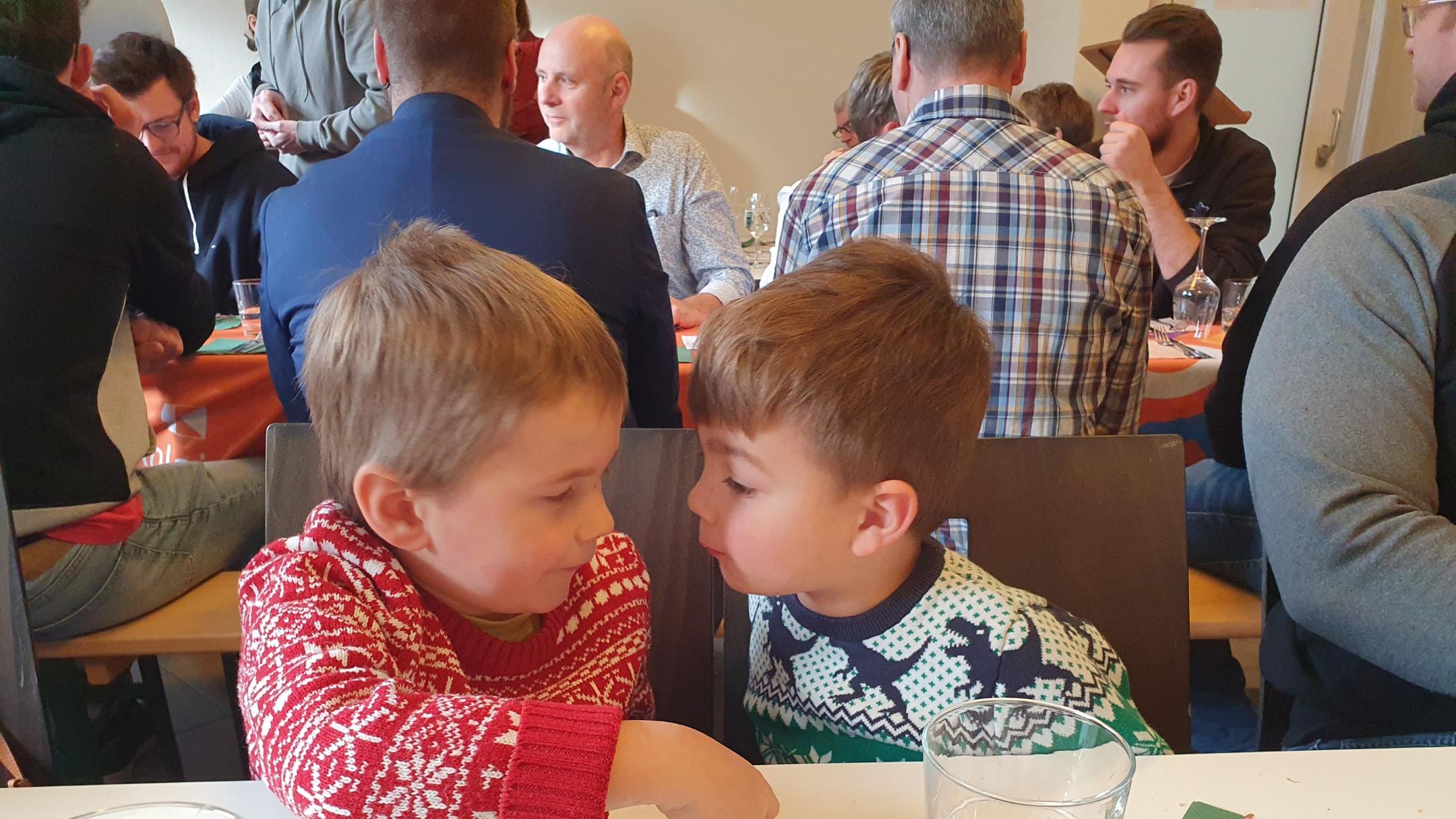 im Vordergrund zwei Kinder in Weihnachtspullis, im Hintergrund Menschen im Gespräch