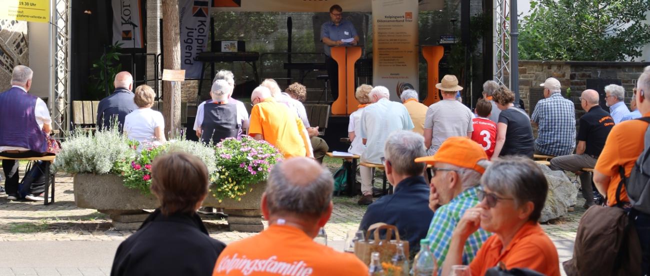 An Tischen und auf Bänken verfolgen Menschen, was auf der Bühne passiert.