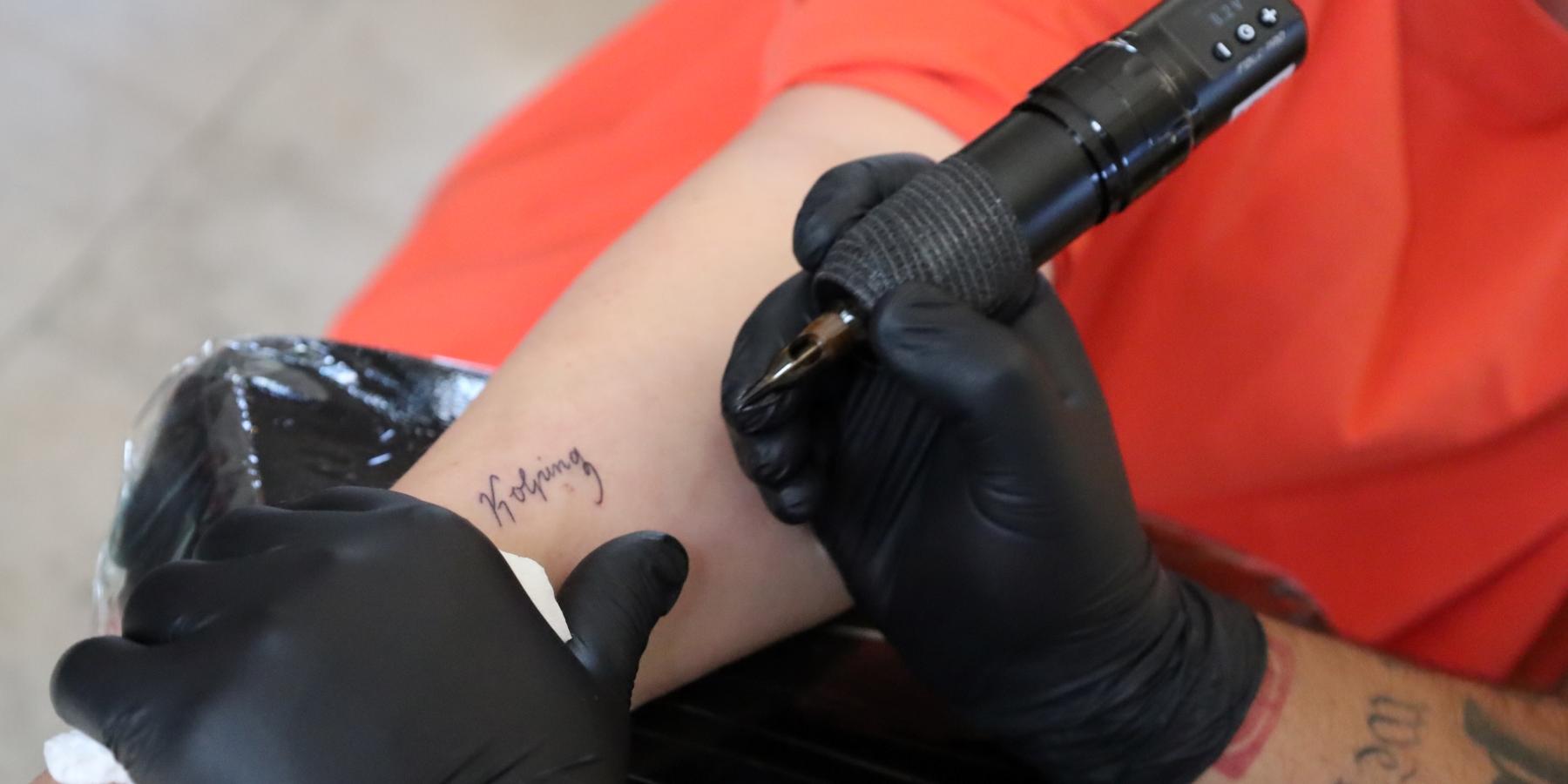 Das Bild zeigt die Hände eines Tattooartisten, der den Schriftzug Kolping auf einen Arm tätowiert.