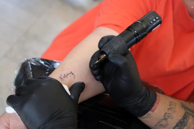 Das Bild zeigt die Hände eines Tattooartisten, der den Schriftzug Kolping auf einen Arm tätowiert.