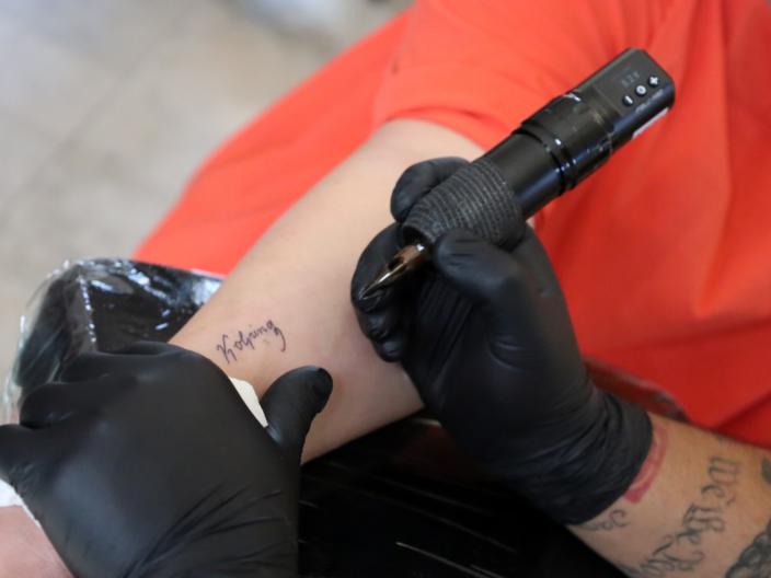 Das Bild zeigt die Hände eines Tattooartisten, der den Schriftzug Kolping auf einen Arm tätowiert.