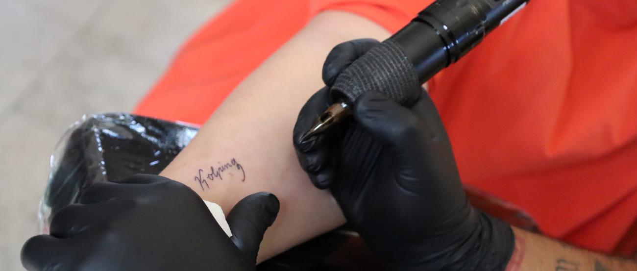 Das Bild zeigt die Hände eines Tattooartisten, der den Schriftzug Kolping auf einen Arm tätowiert.