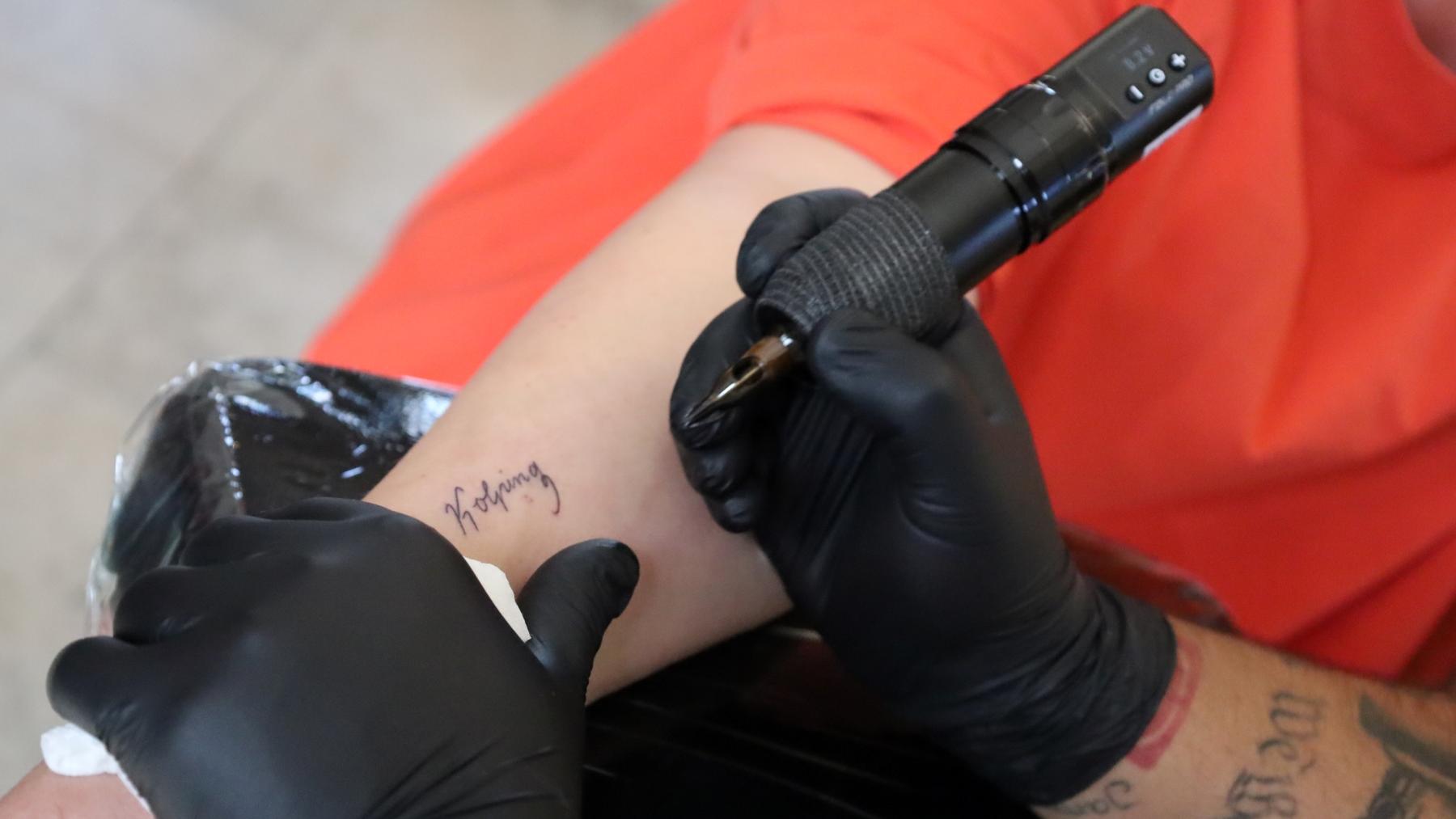 Das Bild zeigt die Hände eines Tattooartisten, der den Schriftzug Kolping auf einen Arm tätowiert.
