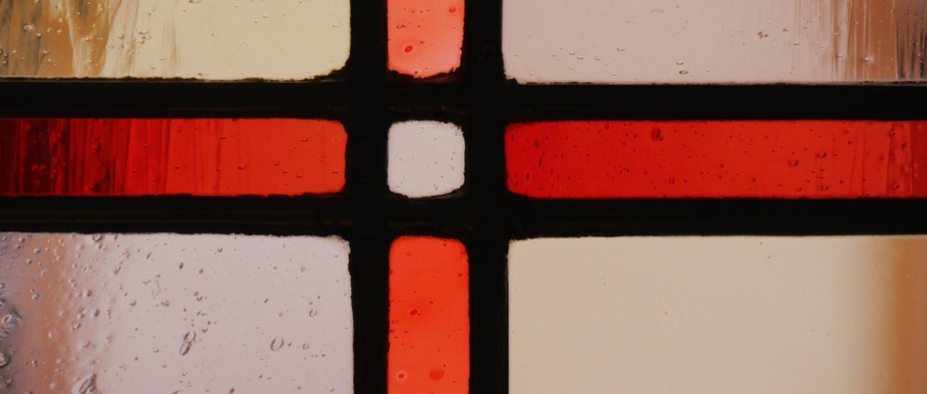 Ausschnitt eines Buntglasfensters mit einem roten Kreuz in durchsichtigen Glas, im Hintergrund scheint die gegenüberliegende Hauswand durch.