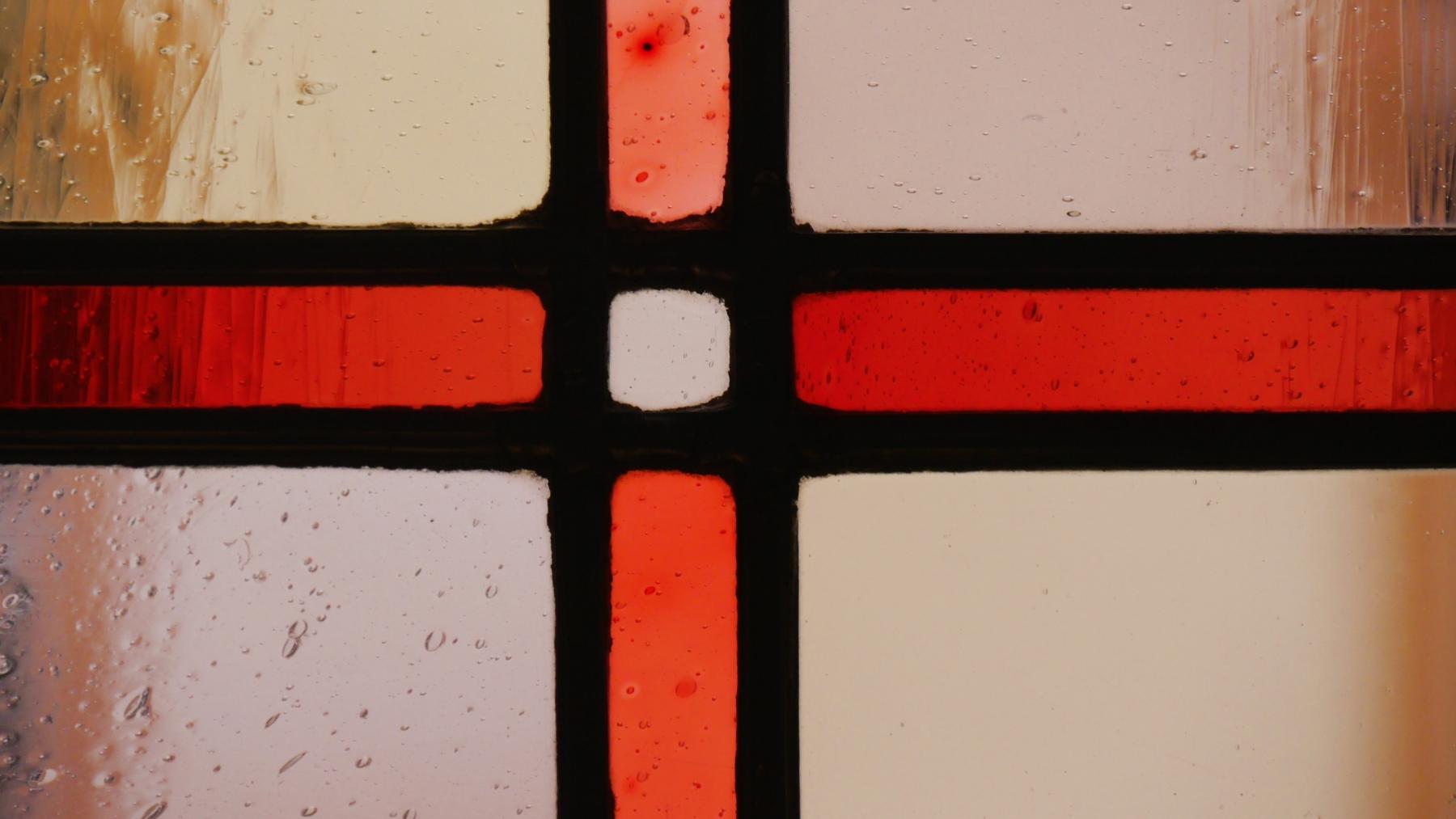 Ausschnitt eines Buntglasfensters mit einem roten Kreuz in durchsichtigen Glas, im Hintergrund scheint die gegenüberliegende Hauswand durch.