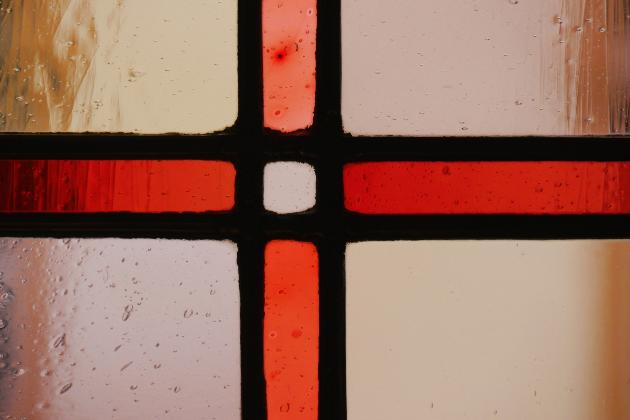 Ausschnitt eines Buntglasfensters mit einem roten Kreuz in durchsichtigen Glas, im Hintergrund scheint die gegenüberliegende Hauswand durch.
