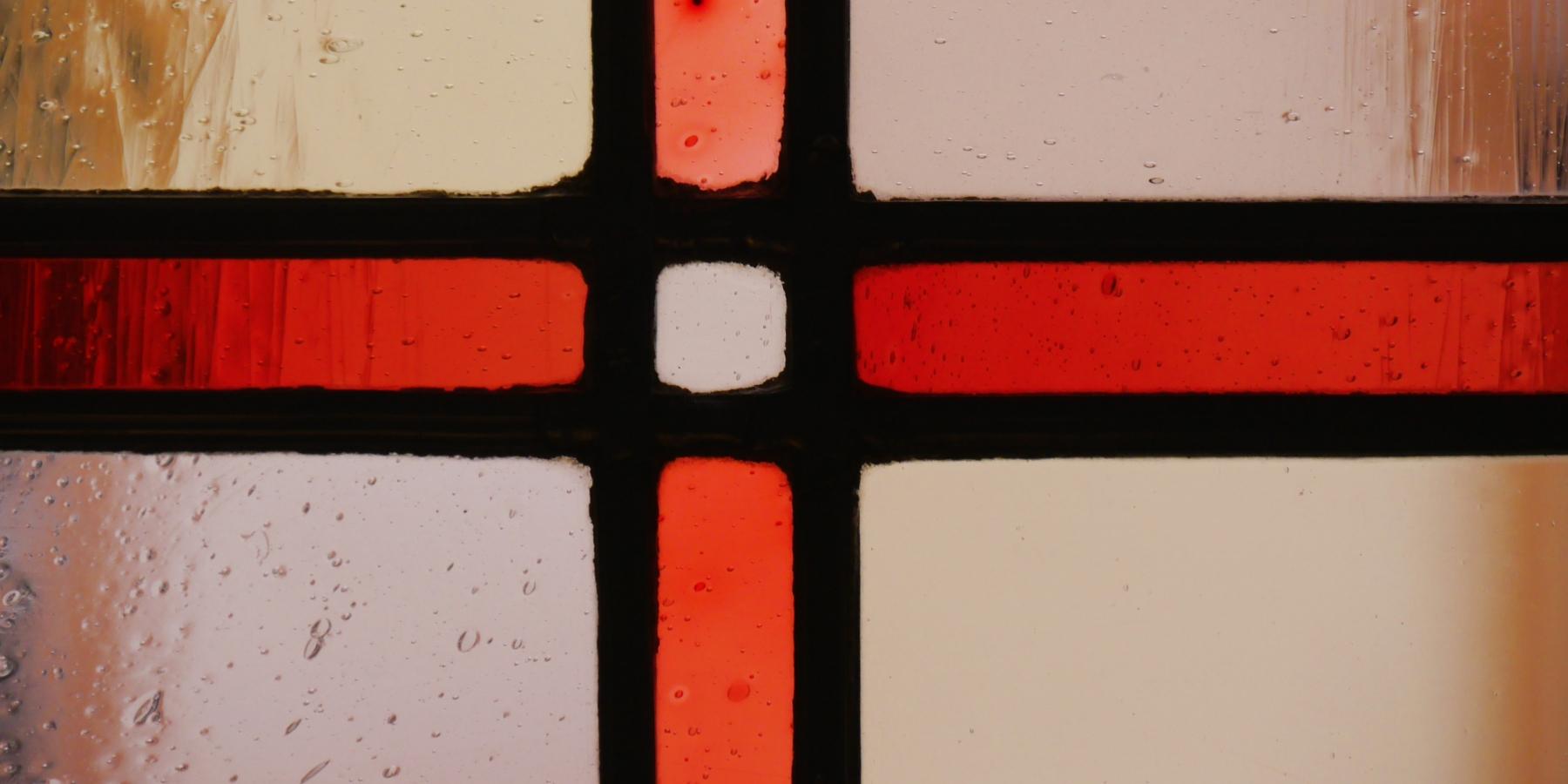 Ausschnitt eines Buntglasfensters mit einem roten Kreuz in durchsichtigen Glas, im Hintergrund scheint die gegenüberliegende Hauswand durch.