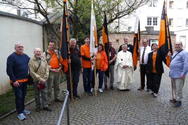 Menschen stehen im Halbkreis im Hof der St. Gangolf-Kirche, einige halten Kolpingbanner.