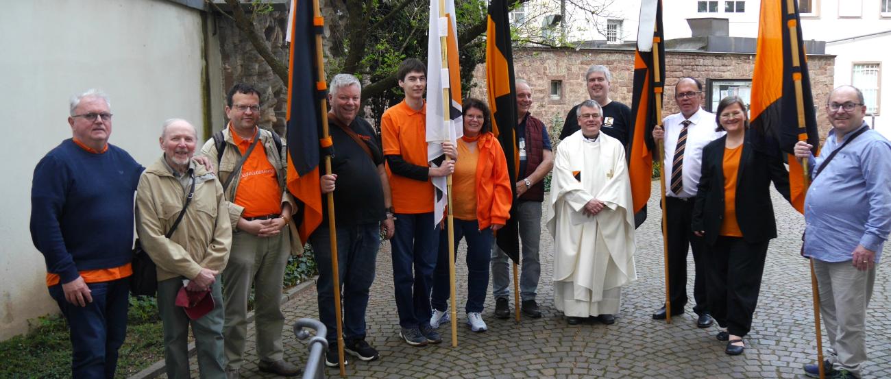 Menschen stehen im Halbkreis im Hof der St. Gangolf-Kirche, einige halten Kolpingbanner.