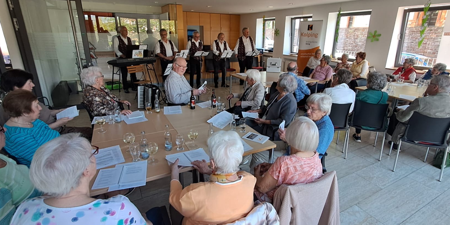 Im Vordergrund sitzende Menschen an Tischen, im Hintergrund die Band 'Hellenbacher Jonge'