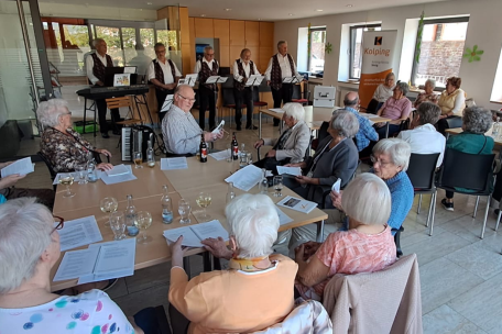 Im Vordergrund sitzende Menschen an Tischen, im Hintergrund die Band 'Hellenbacher Jonge'