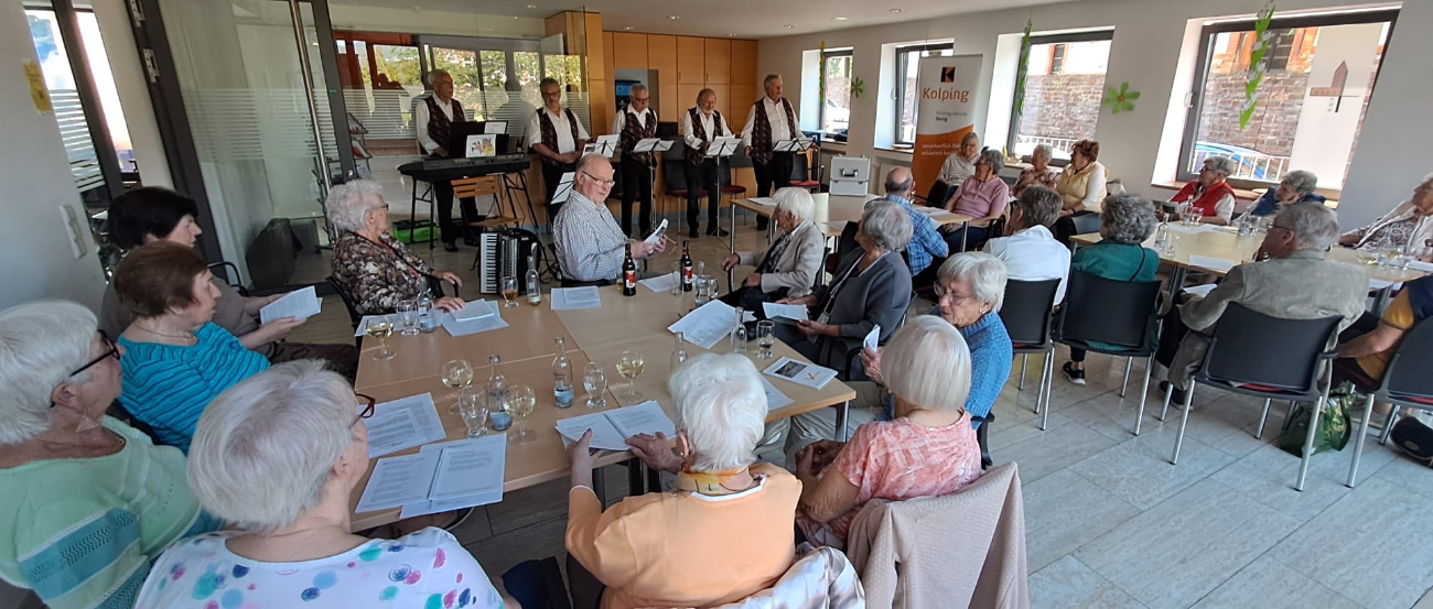 Im Vordergrund sitzende Menschen an Tischen, im Hintergrund die Band 'Hellenbacher Jonge'