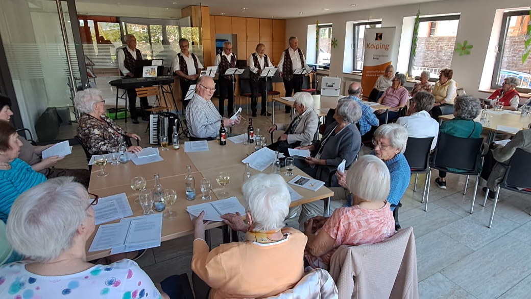 Im Vordergrund sitzende Menschen an Tischen, im Hintergrund die Band 'Hellenbacher Jonge'