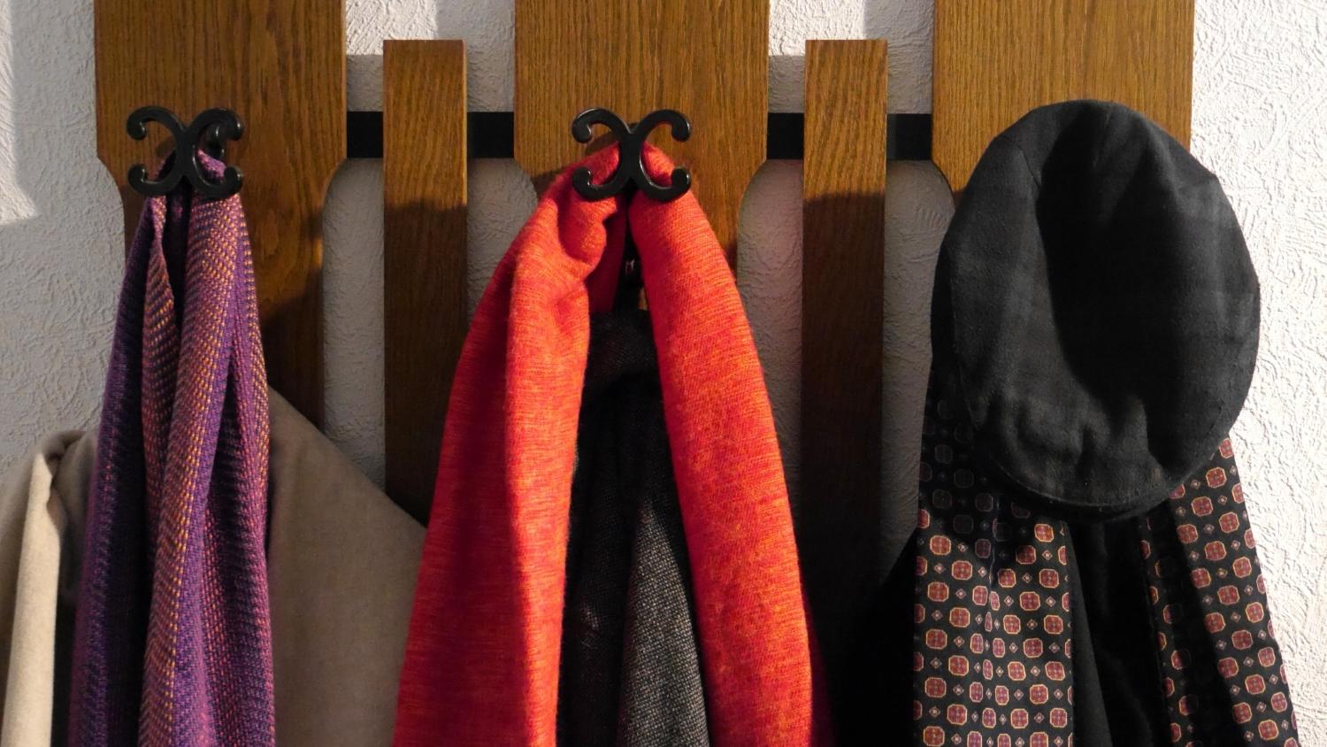 An einer Garderobe hängen drei Wintermäntel mit jeweils einem Schal sowie eine Kappe.