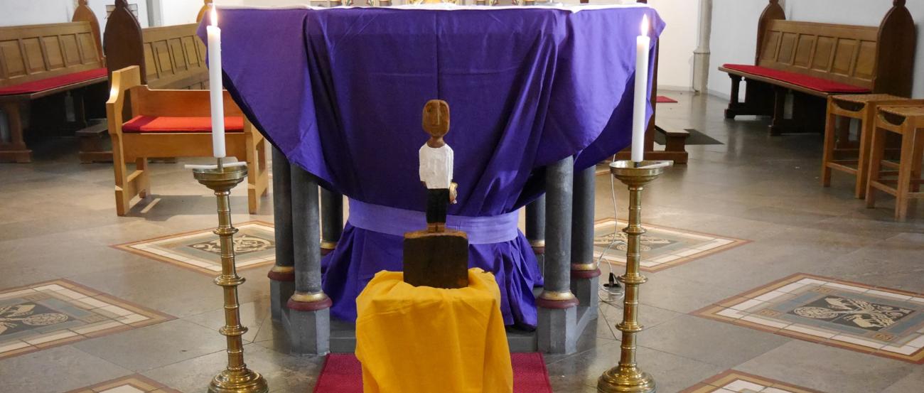 Vor einem Altar steht eine Königsskulptur, davor ein Weg aus Tüchern gelegt mit Steinen, Dornen und welkem Laub darauf, daneben drei Kerzen.