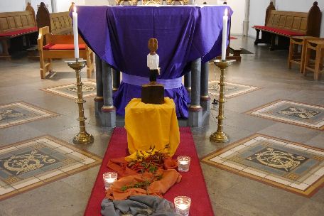 Vor einem Altar steht eine Königsskulptur, davor ein Weg aus Tüchern gelegt mit Steinen, Dornen und welkem Laub darauf, daneben drei Kerzen.