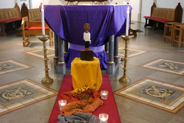 Vor einem Altar steht eine Königsskulptur, davor ein Weg aus Tüchern gelegt mit Steinen, Dornen und welkem Laub darauf, daneben drei Kerzen.