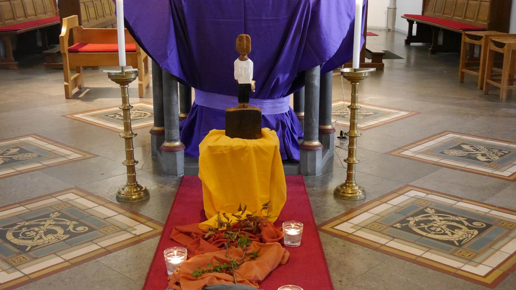 Vor einem Altar steht eine Königsskulptur, davor ein Weg aus Tüchern gelegt mit Steinen, Dornen und welkem Laub darauf, daneben drei Kerzen.