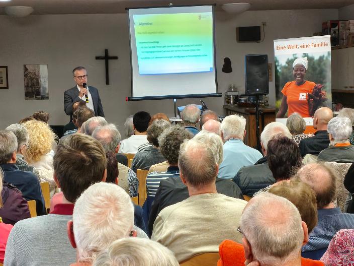 Das Bild zur Infoveranstaltung zeigt das Publikum von hinten, vorne steht der Referent neben einer Leinwand.