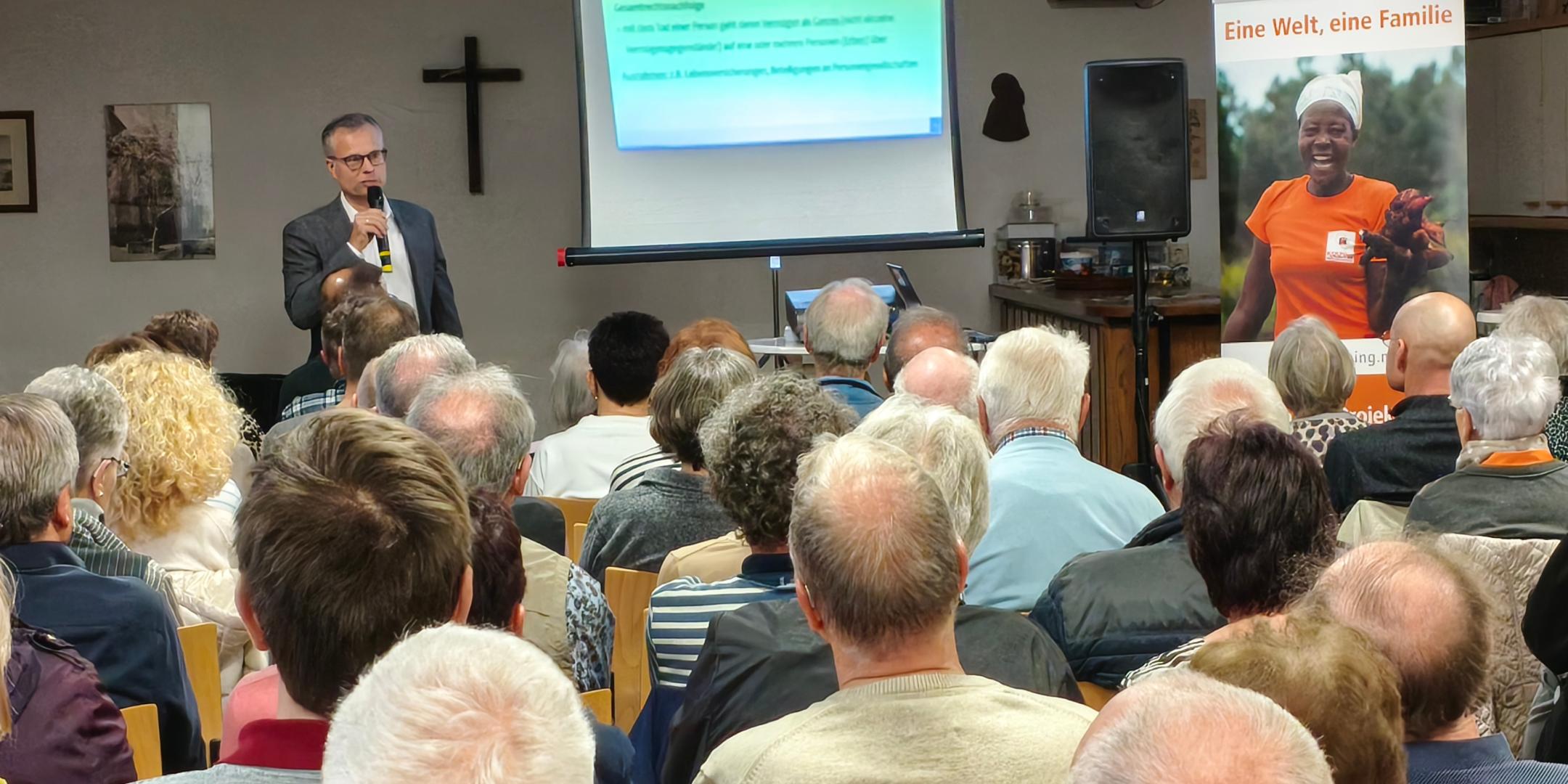 Das Bild zur Infoveranstaltung zeigt das Publikum von hinten, vorne steht der Referent neben einer Leinwand.