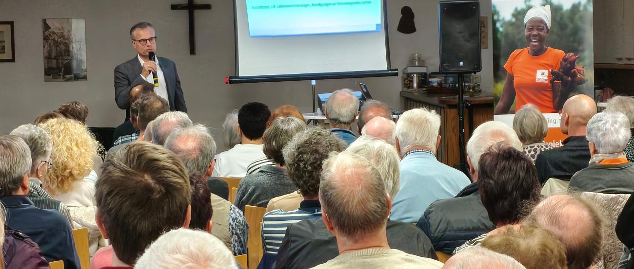 Das Bild zur Infoveranstaltung zeigt das Publikum von hinten, vorne steht der Referent neben einer Leinwand.