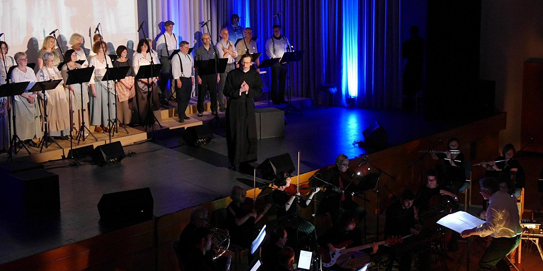 Auf einer Bühne im Scheinwerferlicht ist ein Chor und ein Darsteller in der Rolle des Adolph Kolping zu sehen, vor der Bühne ist ein Orchester.