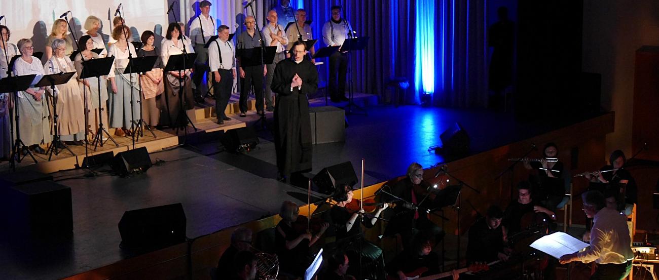 Auf einer Bühne im Scheinwerferlicht ist ein Chor und ein Darsteller in der Rolle des Adolph Kolping zu sehen, vor der Bühne ist ein Orchester.