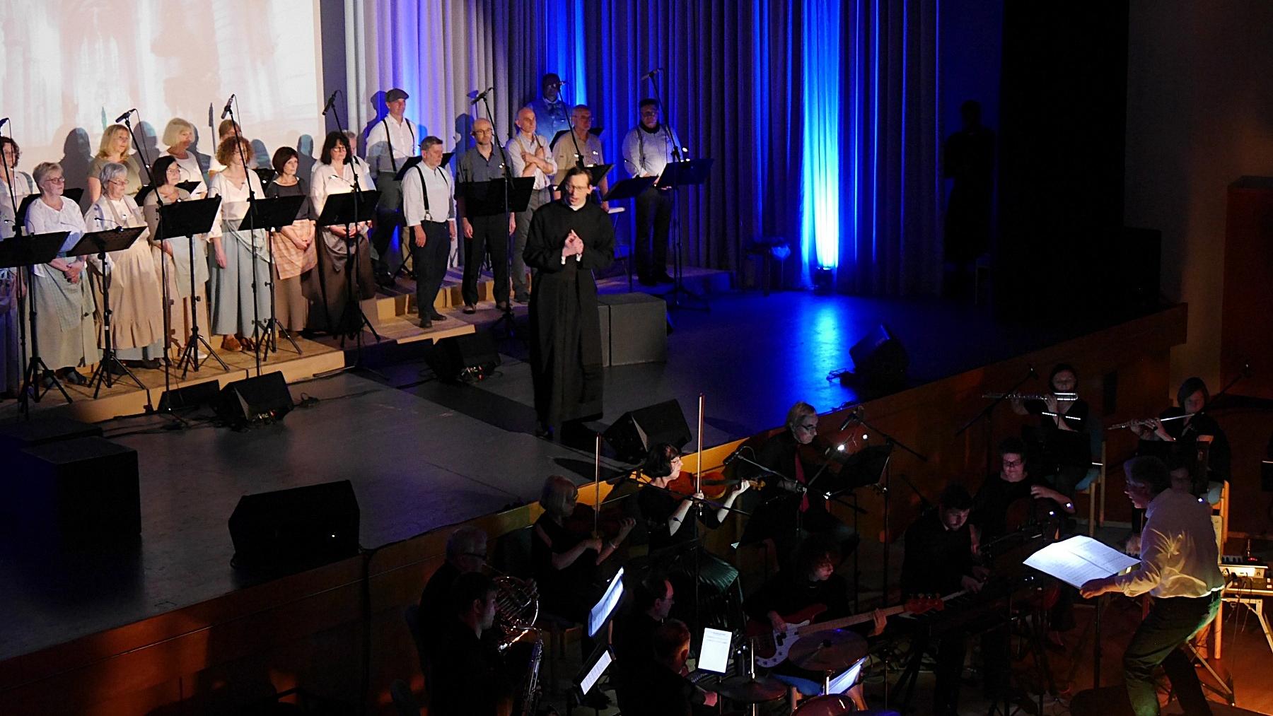 Auf einer Bühne im Scheinwerferlicht ist ein Chor und ein Darsteller in der Rolle des Adolph Kolping zu sehen, vor der Bühne ist ein Orchester.