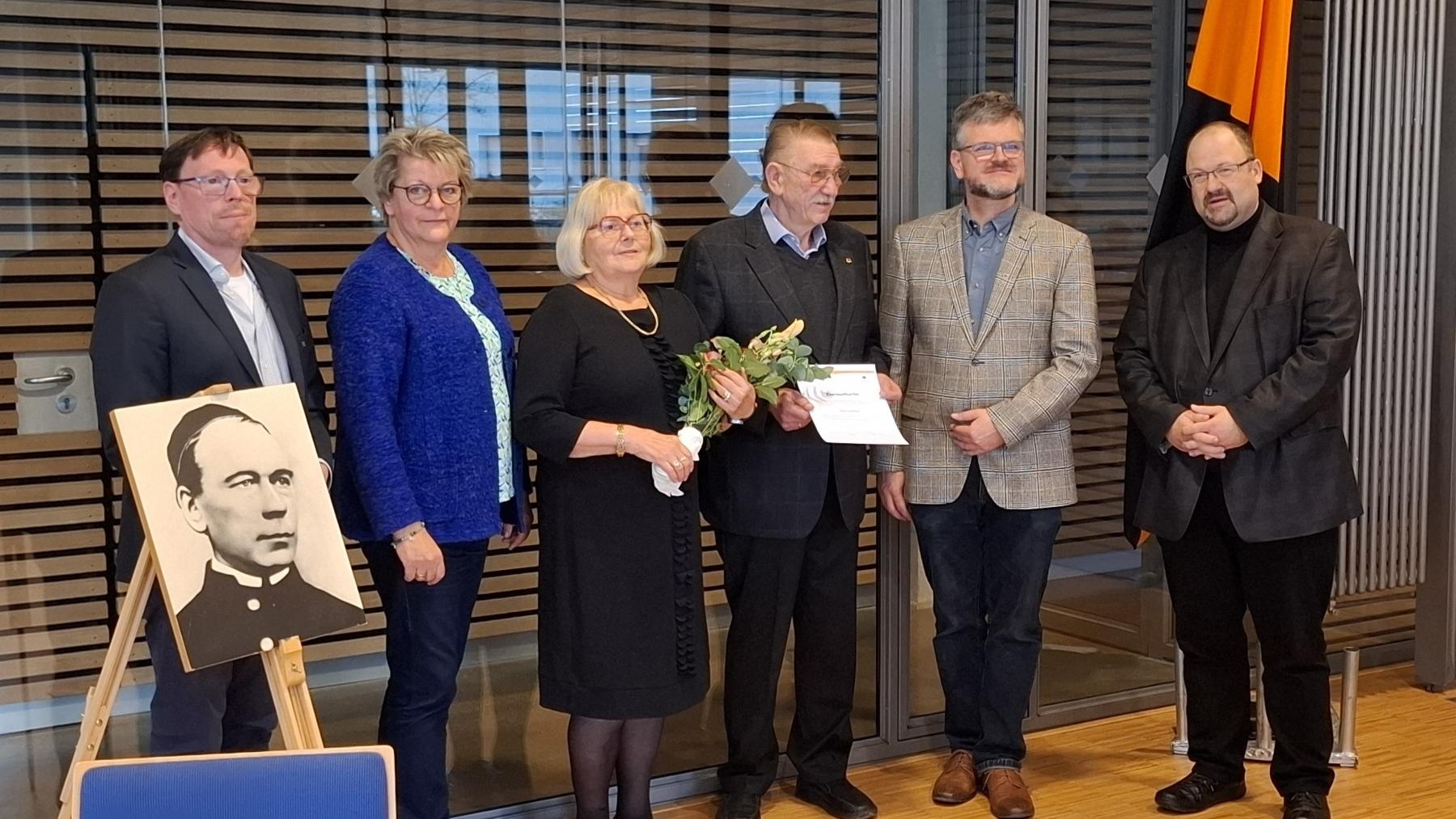 Ehrung des langjährigen Vorsitzenden mit Ehefrau durch das neue Leitungsteam und den Präses, die Personen stehen neben einem Bild von Adolph Kolping.