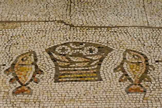 Steinmosaik in Beige- und Brauntönen, das einen Korb mit Broten in der Mitte und an jeder Seite einen Fisch zeigt.
