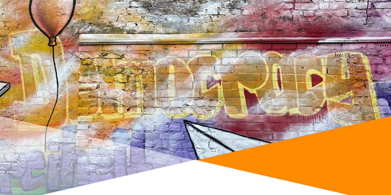 Bild einer bunt besprühten Ziegelmauer mit dem Schriftzug Democracy