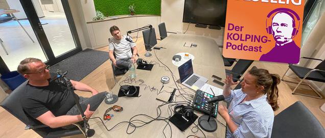 Zwei Männer und eine Frauen sitzen an einem Tisch, es ist viel Aufnahme Technik zu sehen. Sie unterhalten sich