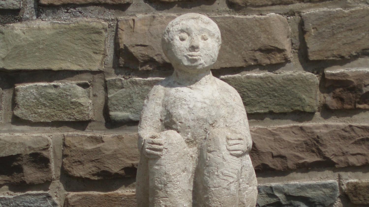 Eine steinerne Figur hält sitzend mit ihren Händen ihre Knie; platziert ist die Statue vor einer Natursteinmauer.