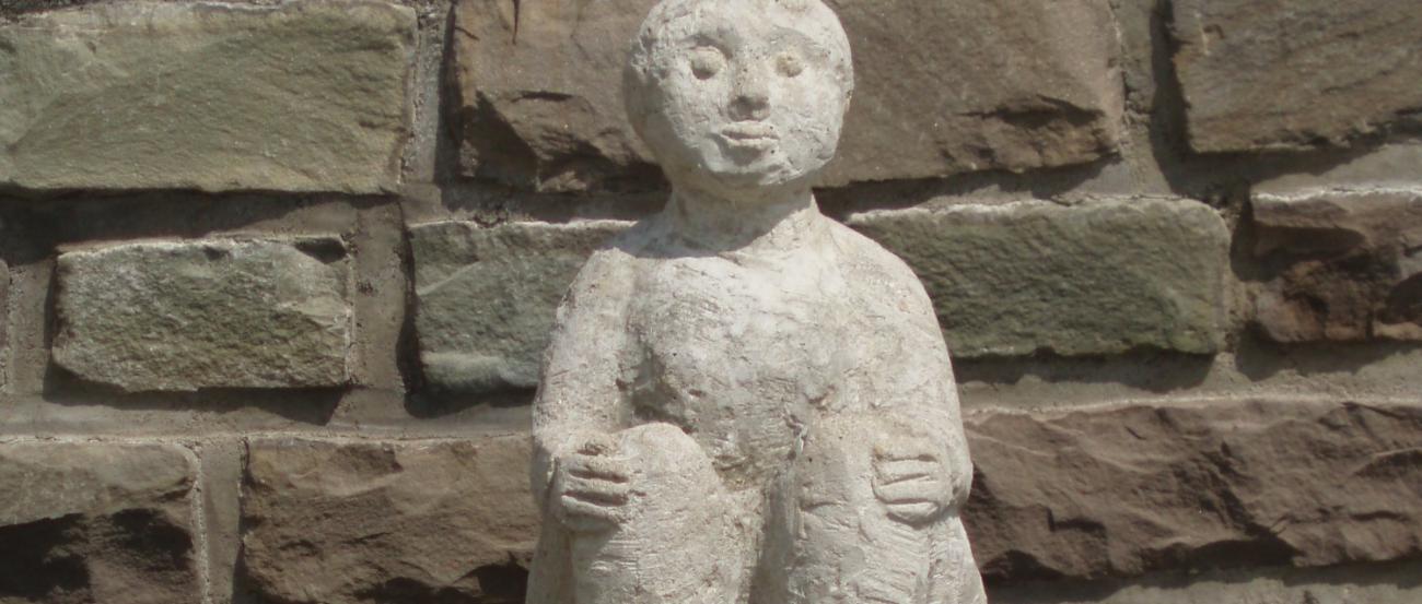 Eine steinerne Figur hält sitzend mit ihren Händen ihre Knie; platziert ist die Statue vor einer Natursteinmauer.