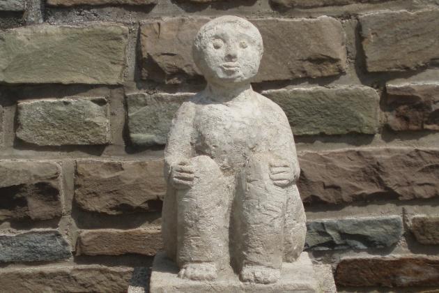 Eine steinerne Figur hält sitzend mit ihren Händen ihre Knie; platziert ist die Statue vor einer Natursteinmauer.