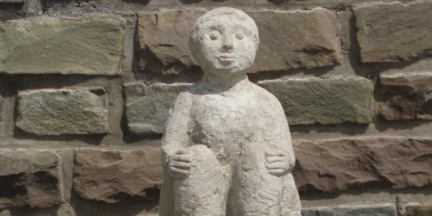 Eine steinerne Figur hält sitzend mit ihren Händen ihre Knie; platziert ist die Statue vor einer Natursteinmauer.