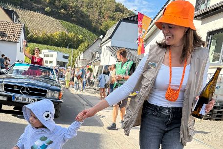 Frau mit orangenem Fischerhut und orangenen Gummistiefeln führt lachend ein Kind an der Hand beim Straßenumzug
