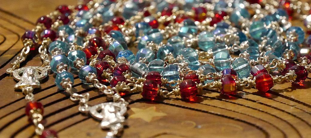 Eine rote und eine blaue Rosenkranz-Kette liegen spiralförmig auf einer Holzunterlage.