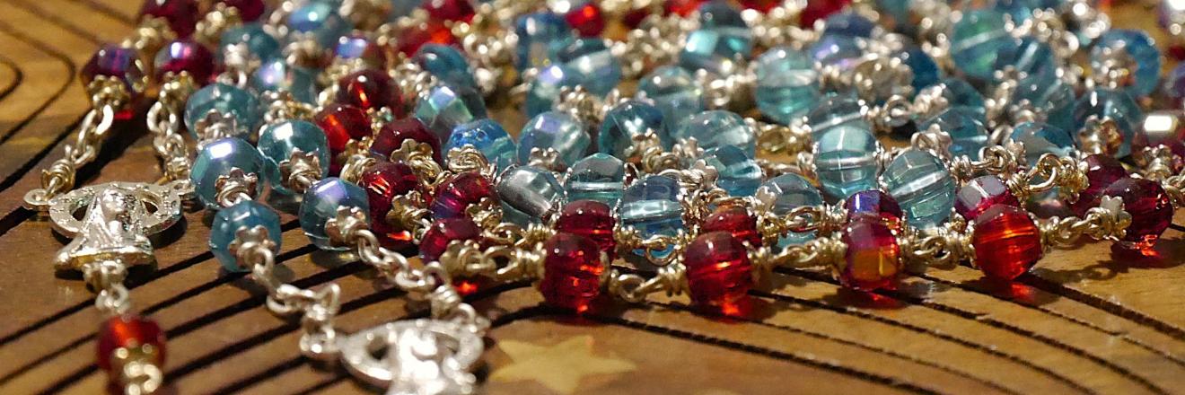 Eine rote und eine blaue Rosenkranz-Kette liegen spiralförmig auf einer Holzunterlage.