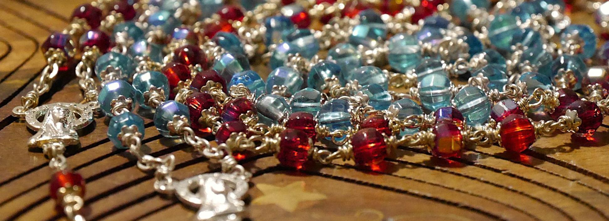 Eine rote und eine blaue Rosenkranz-Kette liegen spiralförmig auf einer Holzunterlage.