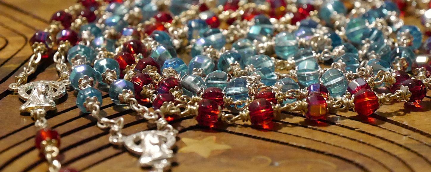 Eine rote und eine blaue Rosenkranz-Kette liegen spiralförmig auf einer Holzunterlage.