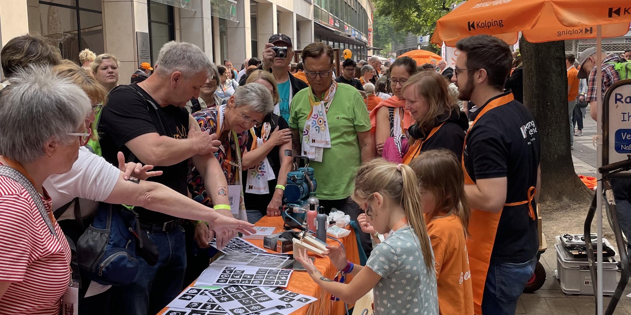 Menschen am orangenen Tisch der Kolpingjugend interessieren sich für die angebotene Aktion 'Airbrush Tattoos'. (c) Kolpingwerk DV Trier Menschen am orangenen Tisch der Kolpingjugend interessieren sich für die angebotene Aktion 'Airbrush Tattoos'.