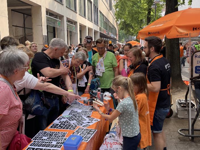 Menschen am orangenen Tisch der Kolpingjugend interessieren sich für die angebotene Aktion 'Airbrush Tattoos'.