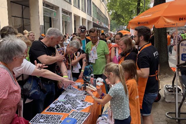 Menschen am orangenen Tisch der Kolpingjugend interessieren sich für die angebotene Aktion 'Airbrush Tattoos'.